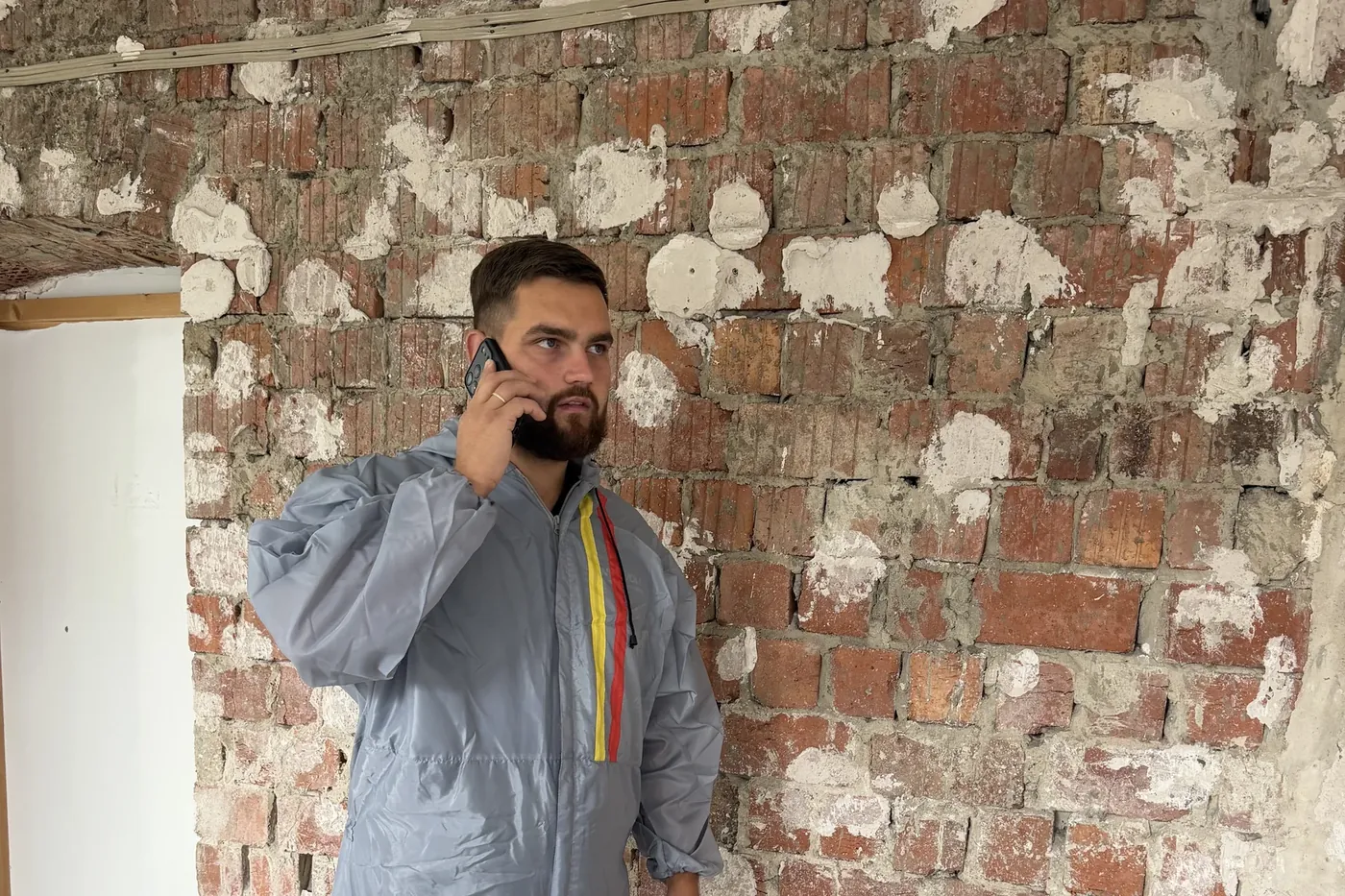 Markus Knaub telefoniert im Schutzanzug vor einer Ziegelwand auf der Baustelle